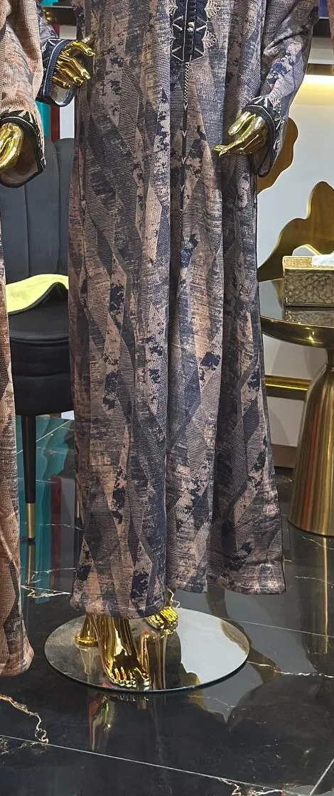 Une djellaba longue à imprimé géométrique dans les tons taupe et bleu nuit, encolure boutonnée, sur un mannequin doré et sans visage.