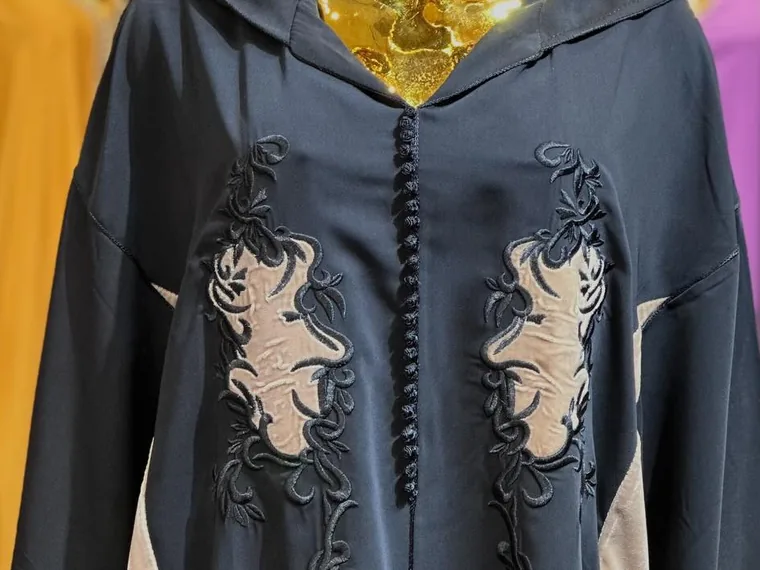 Cadrage rapproché d'une djellaba à capuche bleu nuit&nbsp;: plastron à boutons cordés et appliqués taupe, brodés de rinceaux sombres, sur un mannequin doré.