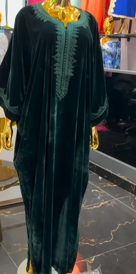 Une gandoura en velours vert foncé, manches chauve-souris, encolure et bords brodés d'arabesques ton sur ton, sur un mannequin doré et sans visage.
