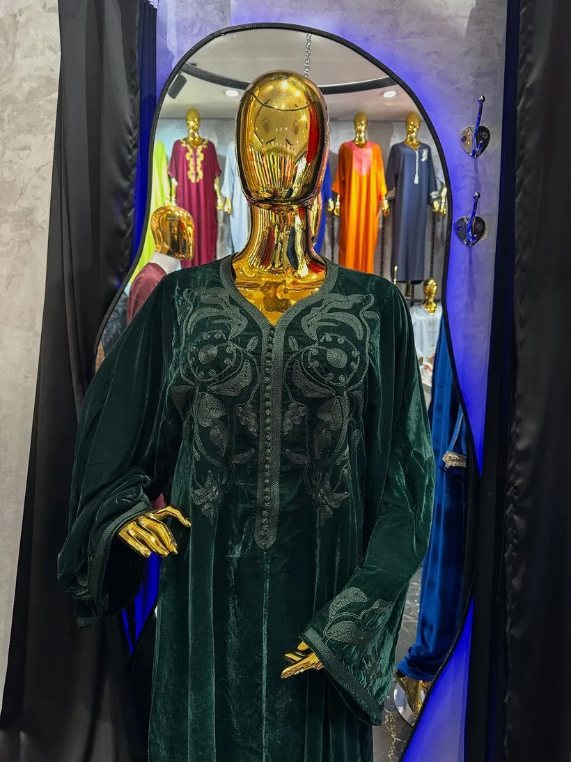 Un caftan en velours vert bouteille, présenté sur un mannequin doré et sans visage, se reflète dans un miroir cintré.